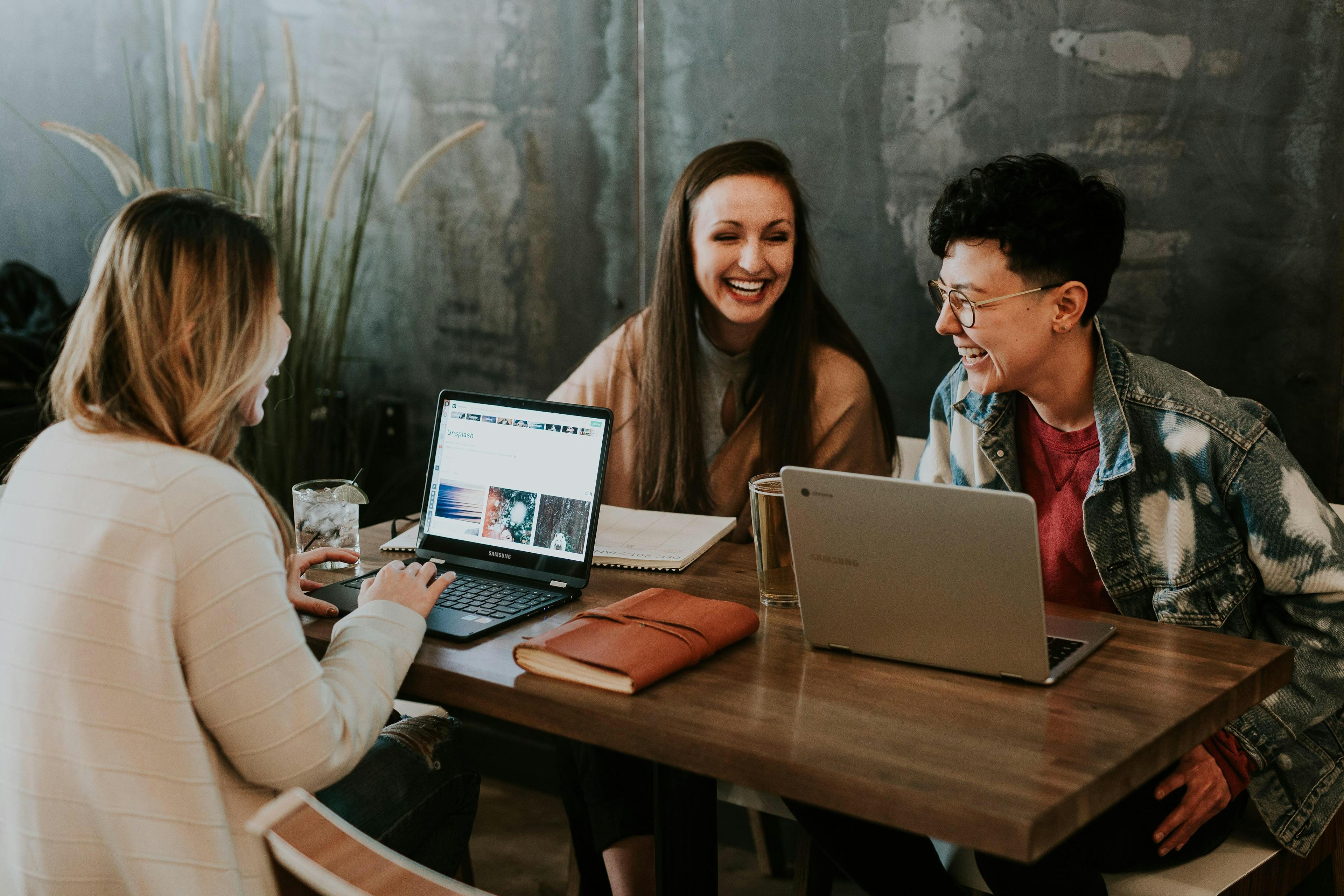 Three people sitting at a table smiling. Two of them have laptops that are opened and one has a notepad in front of her.