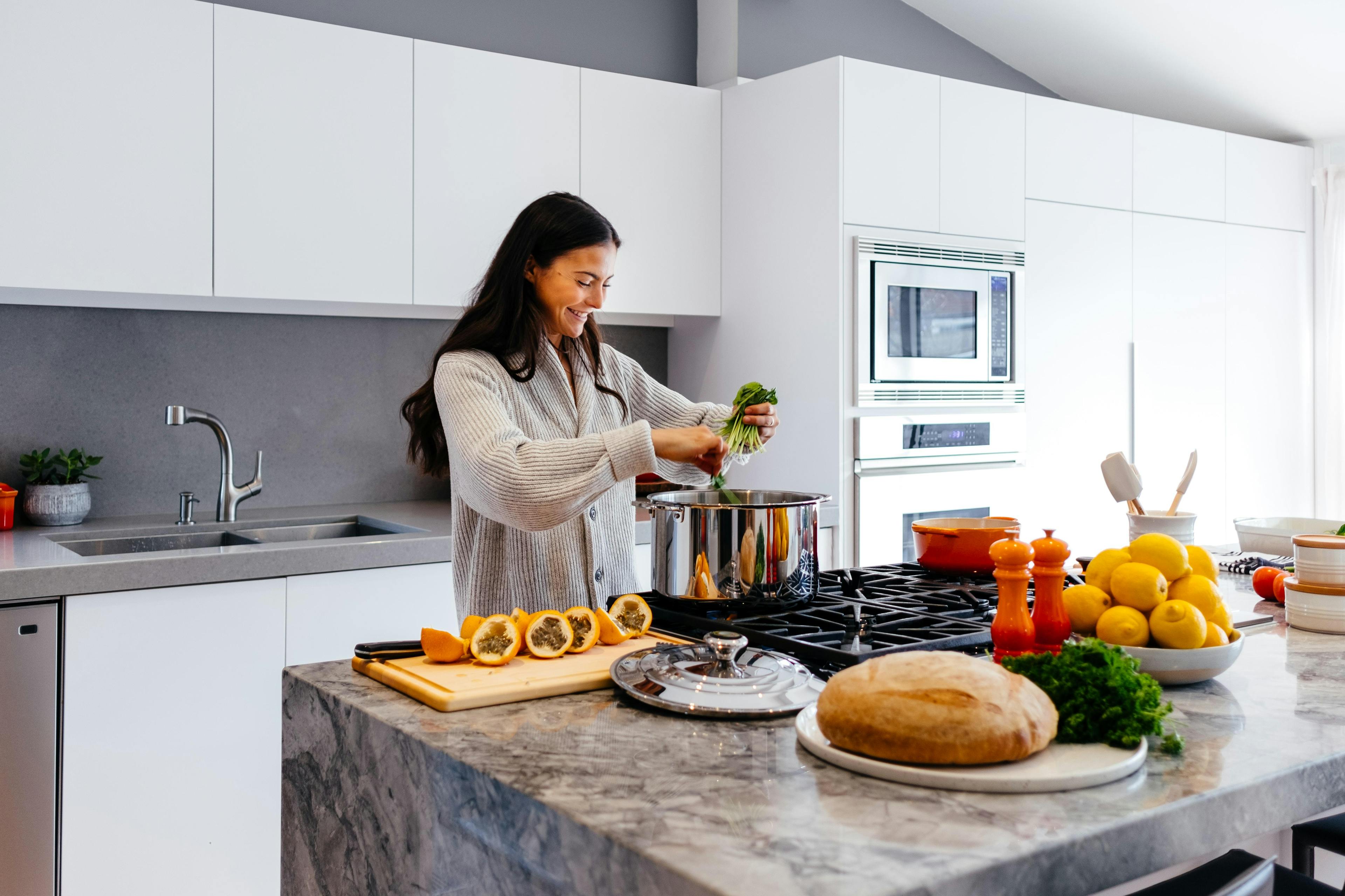 A smiling woman wearing a grey sweater standing in front of a large pot in the kitchen.